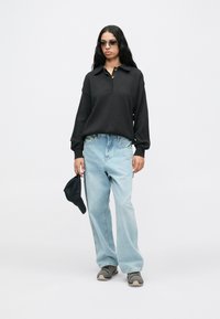 Pessoa jovem a usar uma polo de mangas longas preta, jeans largos azul claros, sapatilhas cinzentas, óculos de sol e segurando um boné preto ao lado.