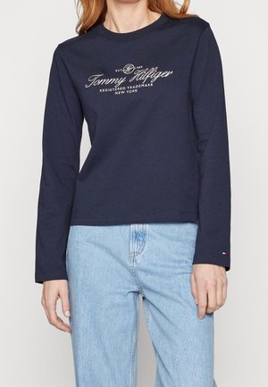 Vrouw draagt een marineblauw Tommy Hilfiger shirt met lange mouwen en witte logotekst, gecombineerd met lichtblauwe jeans, staand tegen een effen achtergrond.