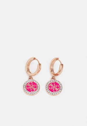 Paire de boucles d'oreilles créoles ton or avec des pendentifs ronds roses présentant un logo doré et une bordure de strass transparents.