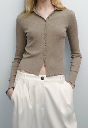 Kvinde iført en beige ribstrikket cardigan med knapper og flæsekant samt højtaljede hvide plisserede bukser, hænderne i lommerne.