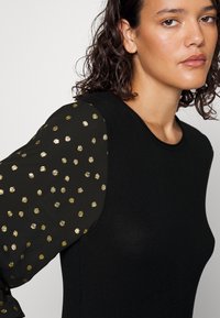 Svart genser med lange, puffete ermer som har gullfargede prikker. Slank, tettsittende design med rund hals og jevn tekstur.