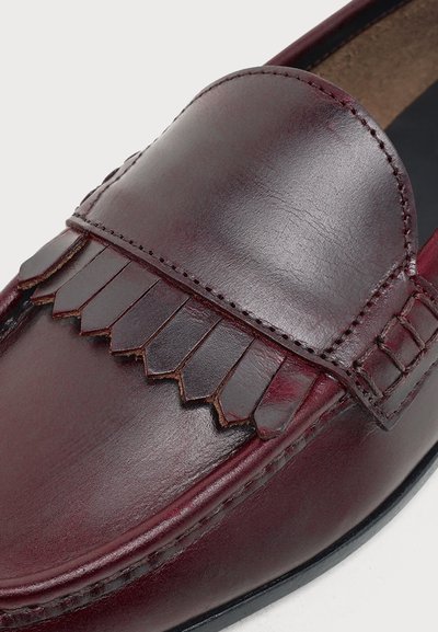 Gros plan d'un mocassin en cuir bordeaux poli avec des franges sur le dessus et des coutures visibles le long des bords.