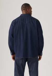 Cămașă din denim închis cu mânecă lungă, cu un design cu yoke ascuțit, mansete cu nasturi și o croială relaxată. Materialul are o textură netedă și o culoare albastră profundă.