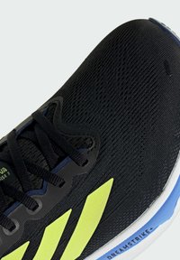 Zapatilla deportiva negra con parte superior de punto texturizado, detalles en amarillo y suela azul en contraste. Cuenta con un sistema de cordones tradicional y marca.