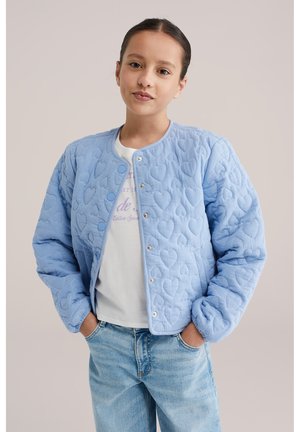 Jong meisje draagt een lichtblauw gewatteerd jasje met hartjespatroon, wit t-shirt met print en lichtblauwe spijkerbroek, handen in de zakken, neutrale achtergrond.