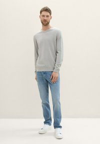 Pull gris clair en V en tissu tricoté doux, associé à un jean bleu clair et des baskets blanches. Le modèle se tient contre un fond neutre.
