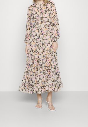 Femme portant une robe longue, fluide, à motif floral avec manches longues et ourlet volanté, associée à des sandales blanches à talons hauts.