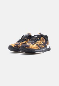 Μαύρα και χρυσά sneakers Versace Jeans Couture με μοτίβο, μαύρα κορδόνια και λευκές λεπτομέρειες στη σόλα, τοποθετημένα δίπλα δίπλα σε λευκό φόντο.