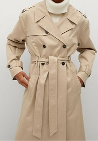 Beżowy płaszcz trench z podwójnym zapięciem na guziki, paskiem w talii i bocznymi kieszeniami. Wykonany z gładkiego materiału o klasycznym designie.