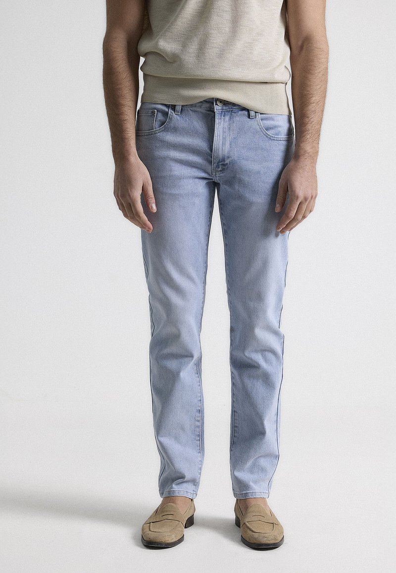 Jeans de denim azul claro con corte recto, bolsillos frontales y un ligero desgaste. Combinados con mocasines color marrón claro y una camiseta sin mangas beige.