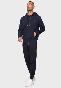 Marineblauwe hoodie en bijpassende joggingsbroek van zacht materiaal. Bevat een oranje trekkoord en zak, met een klein logo op de borst.