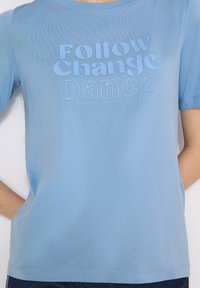 T-shirt bleu clair avec le texte « Follow Change Dance » et un texte plus petit « Vivez votre propre vie, suivez votre propre chemin » sur le devant.