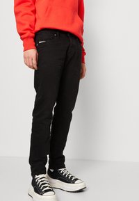 Musta denimmalli, jossa on kapea istuvuus, etutaskut ja vyötäröllä hillitty hopeakoriste, yhdistetty mustavalkoisiin tennareihin.