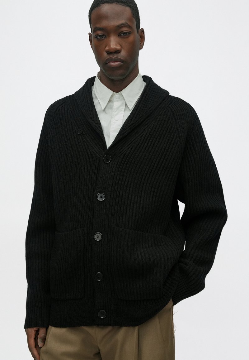 Cardigan nero lavorato a maglia con colletto a scialle, caratterizzato da una texture a costine, apertura con bottoni e due tasche frontali, indossato sopra una camicia chiara.