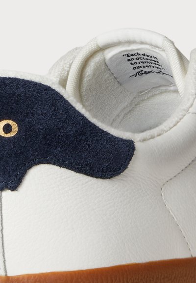 Valge nahast tennisekeng, millel on tumesinised suede aktsendid, ümarad kuldsed silmad, tekstureeritud pind ja pehme vooder. Sisetallal on trükitud tsitaat.