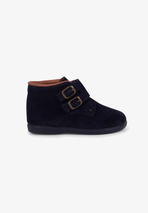 Stivali alla caviglia in camoscio blu navy con doppie fibbie, punta rotonda e fodera interna marrone in contrasto. Suola piatta in gomma con leggera testurizzazione.