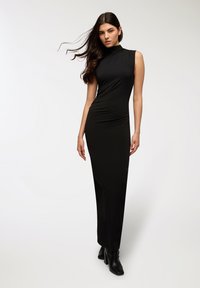 Abito maxi senza maniche nero con collo alto, silhouette aderente e texture liscia, abbinato a stivaletti neri con tacco alla caviglia.