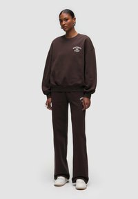 Felpa oversized marrone e pantaloni abbinati, realizzati in morbido tessuto. Caratterizzati da un collo a giro e un logo ricamato in bianco sul petto.