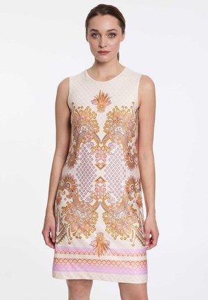 Frau trägt ein ärmelloses, knielanges cremefarbenes Kleid mit kunstvollen symmetrischen floralen und geometrischen Mustern in Orange-, Pink- und Goldtönen.