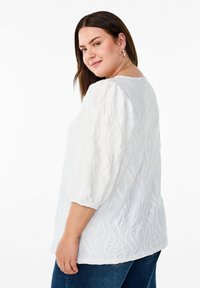 Hvid, struktureret bluse med et bølget mønster, trekvart-pufærmer og rund halsudskæring. Kombination med blå jeans.