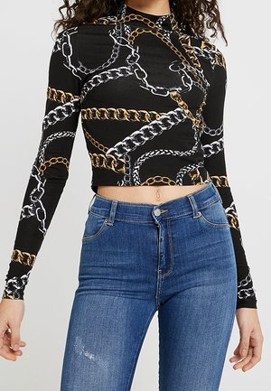Woman wearing a black cropped long-sleeve top with gold and silver chain patterns paired with high-waisted blue jeans.