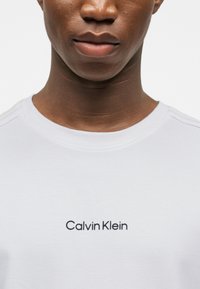 Κοντινό πλάνο ενός ατόμου που φοράει λευκό μπλουζάκι με την επιγραφή "Calvin Klein" σε μαύρο, κεντραρισμένη στο στήθος.