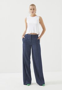 Witte geribbelde mouwloze top met een cropped ontwerp, gecombineerd met hoge taille marineblauwe broek met wijde pijpen, voorzien van witte stippen en groene accenten.