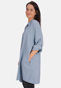 Hellblaues, übergroßes Hemd aus strukturiertem Stoff mit einem Knopfkragen, hochgerollten Ärmeln und seitlichen Taschen.