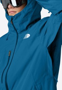 Persona che indossa una giacca impermeabile blu con cappuccio alzato e ventilazione con zip sotto il braccio, mentre sistema il cappuccio vicino agli occhiali da sci.