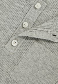 Suéter gris de canalé con cuello, que cuenta con cuatro botones blancos y un bolsillo de parche en el lado izquierdo. Textura suave y diseño casual.