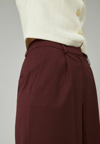 Pantalons ajustés bordeaux avec une texture lisse, présentant des plis avant et des poches latérales, associés à un haut tissé beige clair et des boutons dorés.