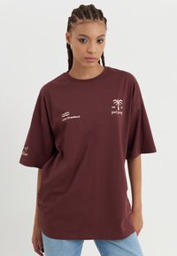 Burgunderfarget oversized t-skjorte laget av bomull, med trykkede detaljer: et palmetre og tekst. Korte ermer og rund hals.