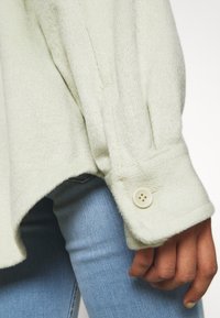 Camicia button-up verde chiaro e texturizzata con orlo arrotondato e un solo bottone sui polsini, abbinata a jeans blu.