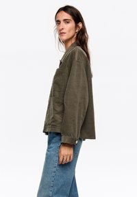Oliwkowo-zielona, fakturowana kurtka z kołnierzem, ozdobiona guzikami i z kieszenią na piersi, zestawiona z jasnoniebieskimi, szerokimi jeansami.