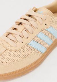 Beige sneakers med vævet tekstiloverdel, lyseblå striber og beige snørebånd. Har gummisål med tekstureret sål for godt greb.