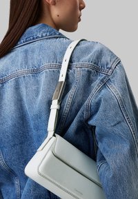 Bolso de cuero blanco con un diseño estructurado, que cuenta con un detalle de hardware en tono plateado, llevado sobre una chaqueta de mezclilla azul.