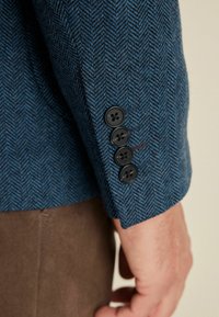 Blaue Stoff mit Fischgrätenmuster und strukturiertem Finish, mit vier schwarzen Knöpfen am Ärmel eines maßgeschneiderten Blazers.