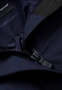 Chaqueta de abrigo navy con una textura suave, que presenta una cremallera negra, etiqueta de tela y un forro interior en contraste. La cremallera está parcialmente abierta.
