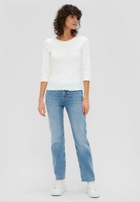 Witte blouse met lange mouwen en een golvende zoom, gecombineerd met lichtblauwe rechtlopende jeans en witte sneakers. Krullend haar zichtbaar.