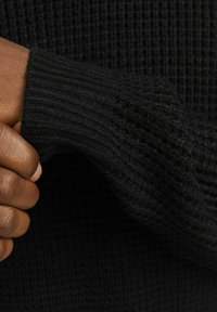 Czarny sweter o fakturowej strukturze z ryżowym splotem. Zbliżenie pokazuje rękaw i dłoń trzymającą materiał, podkreślając jego grubość.