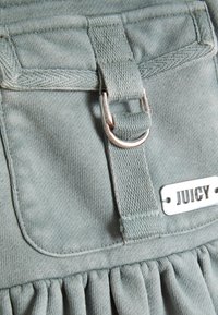 Svetlo modrá tkanina s jemnou textúrou, s striebornou prackou a kovovou štítkom s vyryté "JUICY"; volánkový lem na spodnej strane.