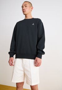 Dunkelgrauer Sweatshirt mit gerippten Bündchen und Saum, mit einem weißen Logo auf der Brust. Kombiniert mit weißen Shorts, die ein einfaches Design zeigen.
