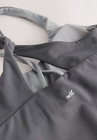 Mørkegrå sports-bh med tynde stropper på en lysegrå baggrund, med hvid tekst "self." på fronten.