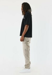 Camiseta negra de manga corta, de corte holgado, combinada con pantalones beige claros que tienen bolsillos. Zapatillas blancas completan el conjunto.