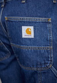 Farkkuhousut, joissa takataskussa valkoinen etiketti keltaisella logolla. Sininen kangas näkyvillä saumoilla ja sileällä pinnalla.