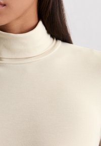 Frau in einem crèmefarbenen gerippten Rollkragenpullover, die Hals, Schulter und einen Teil des Gesichts mit glattem dunklem Haar zeigt.