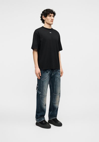 Άνδρας που στέκεται φορώντας μαύρο υπερμεγέθες T-shirt με το κείμενο "OFF", ξεβαμμένο μπλε τζιν πατς και μαύρα αθλητικά παπούτσια σε απλό γκρι φόντο.