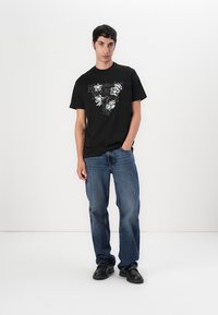 Black cotton t-shirt with floral graphic in white, paired with blue denim jeans. Model wearing dark shoes, standing against a plain background.