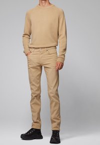 Beige pletený svetr s ribovaným výstřihem, doplněný o béžové slim-fit kalhoty. Oblečení je dokončeno černými šněrovacími botami.