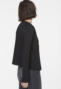 Black jacket with a relaxed fit, long sleeves, and frayed edges. Material appears soft, with side pockets and no visible fastenings.